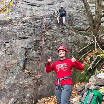 一名学生在山腰前竖起大拇指，表示另一名学生正在攀岩